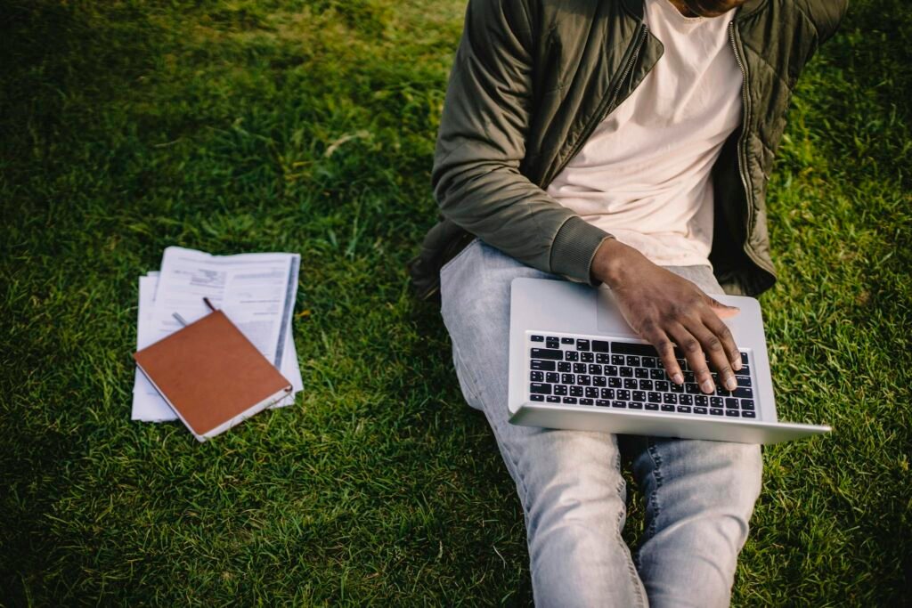 Young adult studying on a laptop outdoors, sitting on grass with documents.